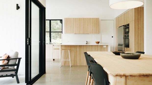 Bright open-plan kitchen and dining area with light timber cabinetry, a long timber dining table, black dining chairs, white walls, pale tiled flooring, and large black-framed glazed doors. A compact living or seating area is visible at the left edge.