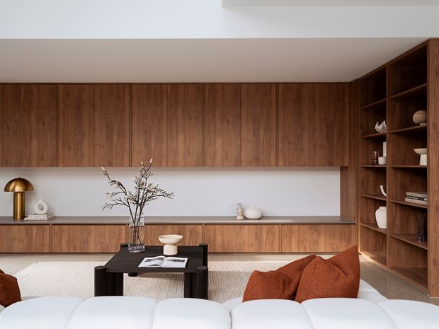 A warm, refined living room viewed from behind a white sofa, featuring extensive dark timber built-in cabinetry across the rear wall and a full-height shelving unit on the right. A low black coffee table, neutral rug, sculptural ceramics, dried branches, and rust-coloured cushions create a contemporary, minimalist interior with strong natural material warmth.