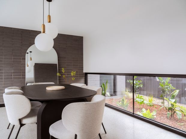 Contemporary dining area with a dark oval dining table surrounded by upholstered cream dining chairs. A large arched mirror sits against a dark textured tiled feature wall, reflecting the sculptural white globe pendant lights. Full-height corner glazing overlooks a landscaped courtyard, while a light polished floor and pale walls keep the space bright and refined.