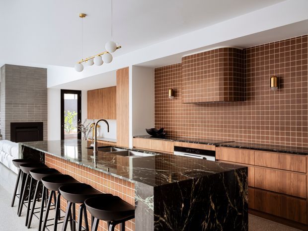 A sophisticated open-plan kitchen with a large island clad in dark veined stone and a tiled waterfall end, paired with a warm timber kitchen and a curved tiled rangehood feature wall. The island includes an undermount sink, sculptural tap and seating for several people on black stools. A living area with a fireplace is visible beyond, while brass and white globe pendant lighting and wall sconces add refined detailing.