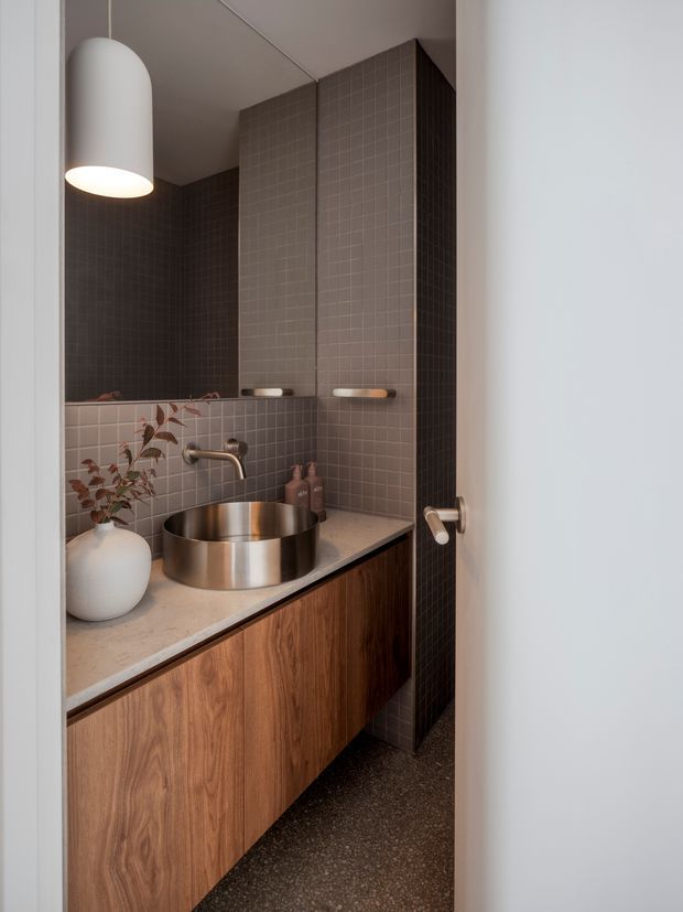 Compact, refined bathroom viewed through an open doorway. The room features dark taupe small-format tiled walls, a floating timber vanity with a pale stone benchtop, a round brushed-metal vessel basin, wall-mounted tapware, integrated shelving, and a large cylindrical pendant light. Warm timber and metal accents contrast with the muted dark tile palette.