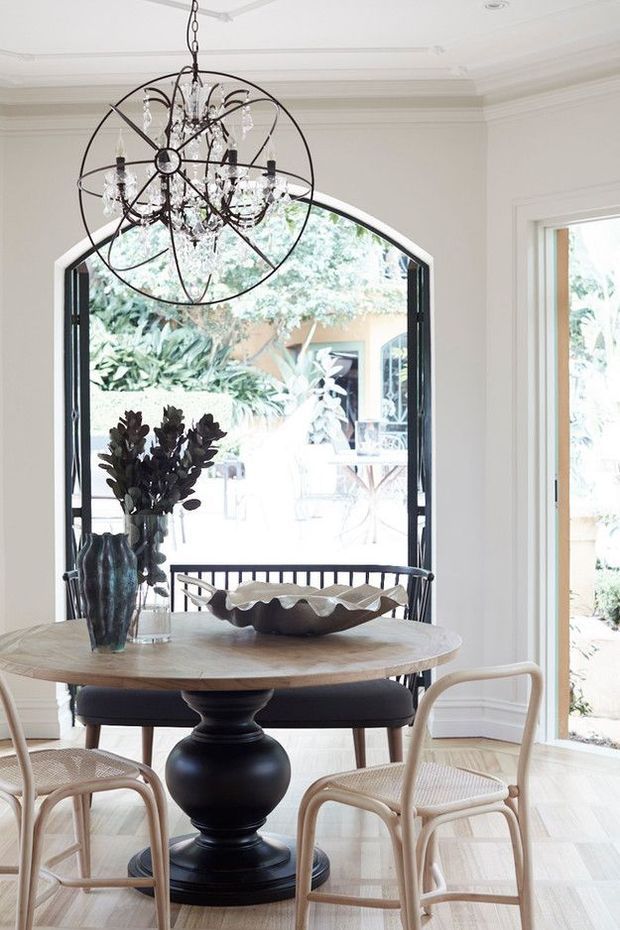 Bright, elegant dining room with a round timber dining table, black pedestal base, woven cane dining chairs, a dark bench seat, and a sculptural black-and-crystal chandelier. The room features white decorative wall mouldings and ceiling cornices, pale timber parquet flooring, and a tall arched glazed opening connecting to an outdoor courtyard or garden.
