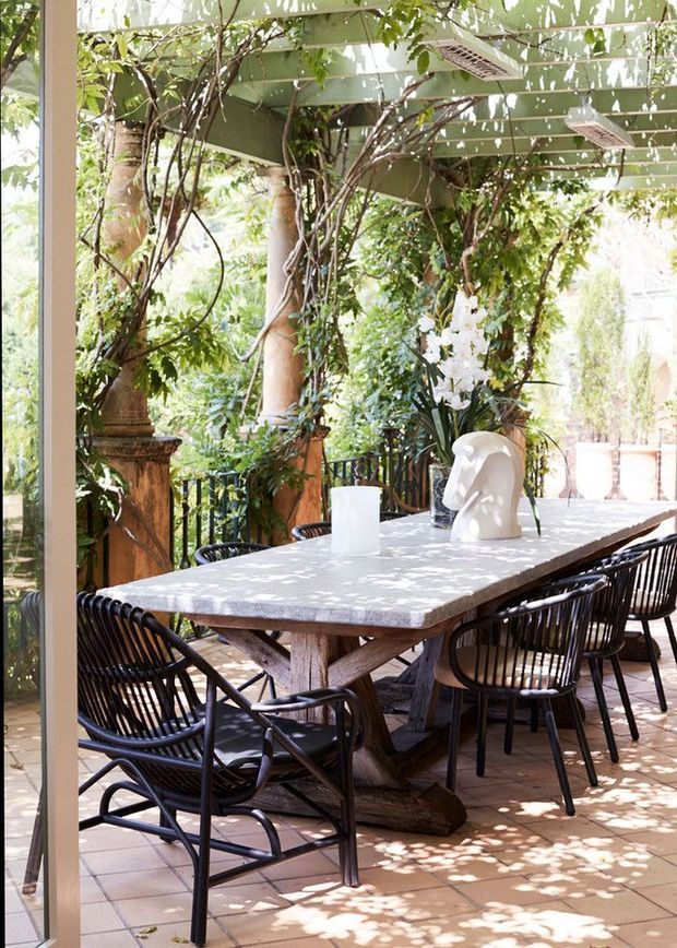 A bright, leafy outdoor dining terrace viewed through a glazed opening, with a large rustic timber dining table, black woven chairs, tiled flooring, and abundant climbing greenery surrounding the space. The terrace has a relaxed, organic character with dappled sunlight and integrated overhead outdoor heaters.