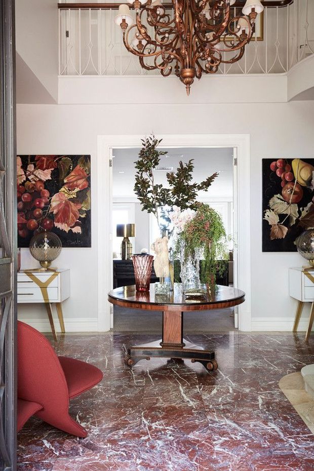 A refined, traditionally styled foyer or entry hall with a dramatic double-height volume and mezzanine balustrade. The room features a highly ornate bronze chandelier, white paneled walls and trim, a round dark-wood pedestal table with floral arrangements, marble flooring in rich burgundy and white veining, and matching botanical artwork and console tables. A seating area is partially visible in the foreground, while a bright adjoining room is seen through the central doorway.