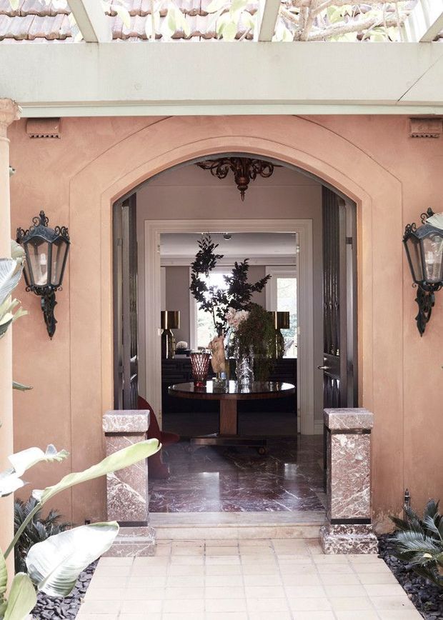 A formal residential entryway viewed from outside, featuring a salmon-pink stucco facade with a large arched opening, ornate black wall lanterns, marble-clad gate pillars, and pale tiled paving. Through the open double doors is a sophisticated interior foyer or living space with dark polished marble flooring, a round console table, floral arrangements, and traditional decorative detailing. A glazed overhead canopy admits bright natural light.