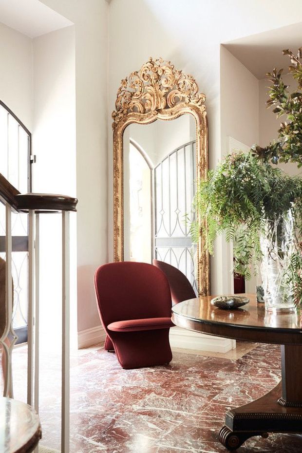 Elegant, light-filled interior featuring an ornate gilded floor mirror, deep burgundy sculptural armchairs, a dark wood console table, and a large indoor fern arrangement. The room has polished reddish marble flooring, pale walls, tall arched glazing, and refined decorative detailing.