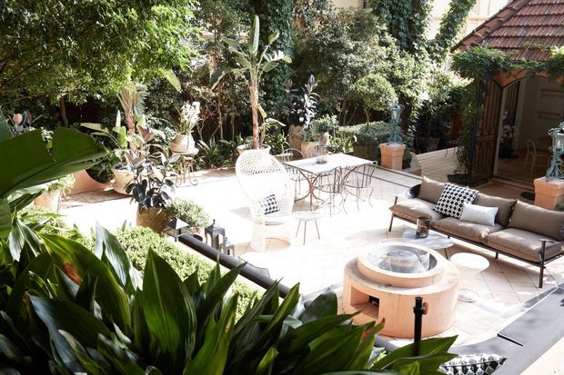A bright, lushly planted outdoor courtyard patio furnished with a low outdoor sofa, armchairs, small tables, and a circular raised fire or water feature. Pale tiled paving, terracotta planters, dense tropical greenery, and a tiled roof structure create a resort-like garden setting.