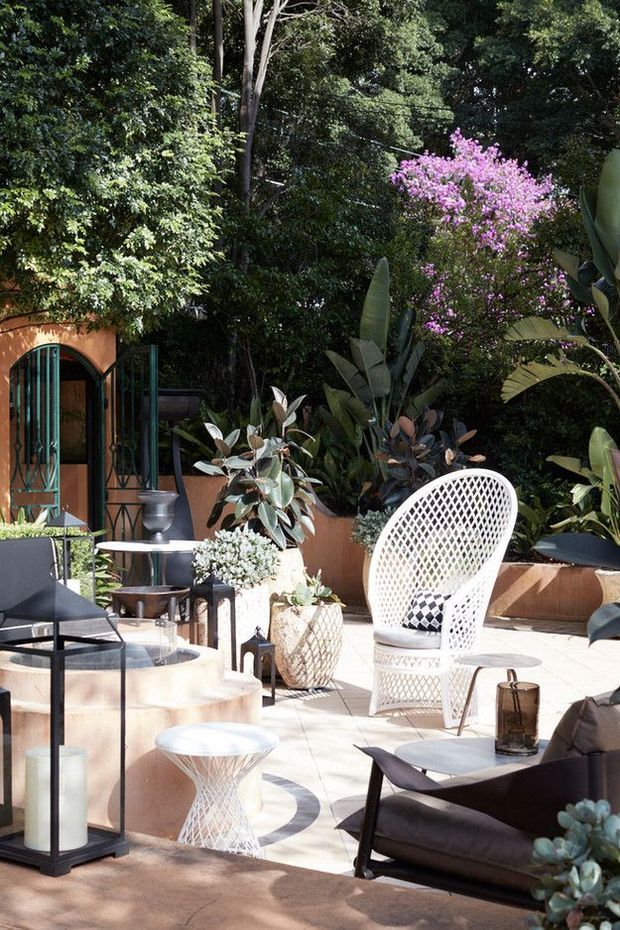 A sunlit landscaped courtyard or terrace furnished as an outdoor lounge. The space features pale tiled or rendered paving, a curved raised planter or seating edge, abundant tropical and leafy planting, a purple flowering tree, a white woven lounge chair, dark contemporary outdoor chairs and tables, lanterns, and decorative potted plants. An arched glazed doorway opens into a terracotta-toned building at the rear.