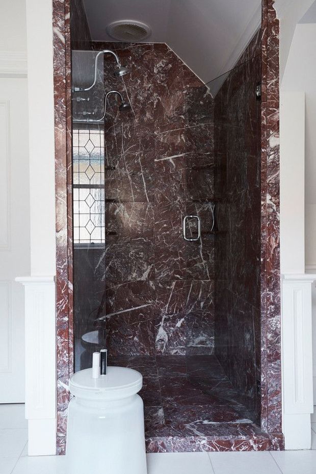 A compact, dramatic shower room clad almost entirely in dark burgundy-red veined marble, including the walls, shower floor, and doorway surround. The space features a rainfall shower, handheld shower fittings, chrome hardware, a narrow leaded-glass window, and a white pedestal toilet in the foreground. White paneled walls and trim frame the richly patterned stone enclosure.