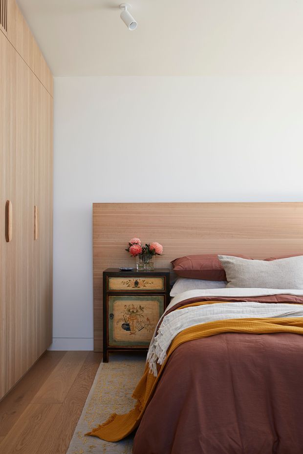 A warm, compact bedroom with a built-in light timber wardrobe along the left wall, matching timber floor and a simple timber headboard. The bed is dressed in muted rust, mustard and cream layered bedding, beside a small vintage-style bedside cabinet with flowers. White walls and ceiling, minimal white track lighting, and soft natural tones create a calm contemporary interior.