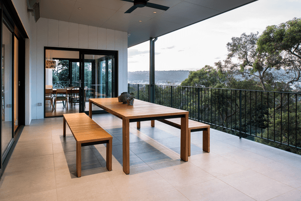 A spacious covered outdoor terrace with a timber dining table and benches, tiled flooring, black metal balustrade, ceiling fan, and expansive views over a wooded landscape and distant water or hills. Glazed doors connect the terrace to an interior dining area.
