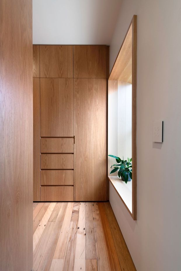 A narrow, light-filled interior passage or entry area with full-height custom timber cabinetry, integrated drawers, warm timber flooring, and a deep timber-framed recessed window seat containing a potted plant. The minimalist joinery and natural wood create a calm, refined atmosphere.