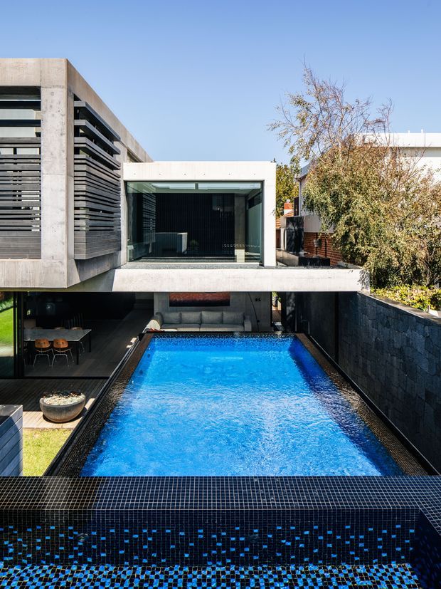 A contemporary multi-level urban residence organized around a long rectangular swimming pool. The image looks toward a glazed upper-level room and concrete-framed architecture, with dark horizontal screening and cladding, a partly visible outdoor dining area, and a tiled pool edge and water feature in the foreground. The design has strong geometric lines, a dark monochromatic palette, and abundant clear blue sky.