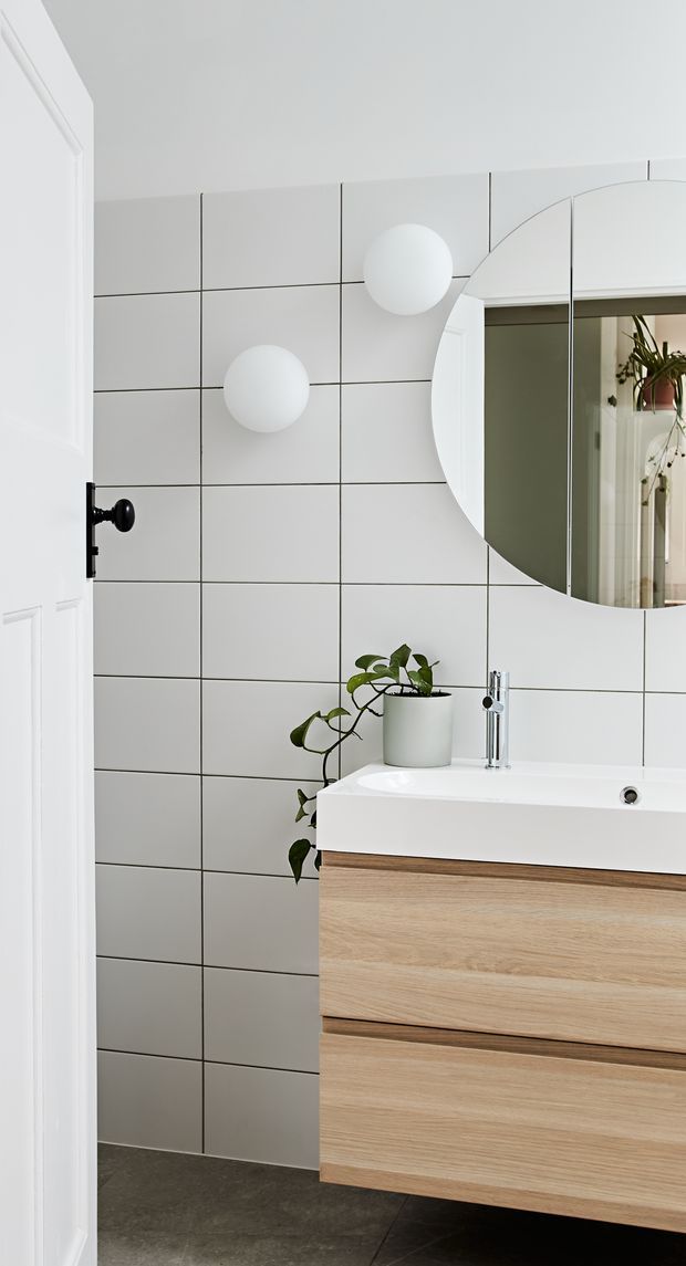 A bright, compact contemporary bathroom with a floating light timber vanity, white integrated basin, chrome wall-mounted tap, large round mirror, and two spherical wall lights. The walls are clad in large white square tiles with dark grout, while the floor is a dark grey finish. A potted trailing plant adds a natural accent, and a white paneled door is visible at the left.