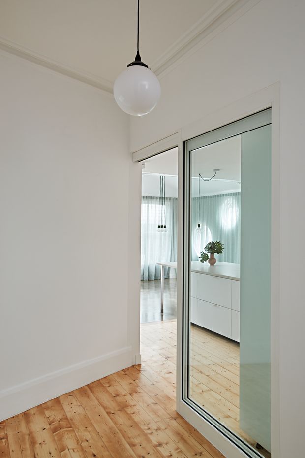 A bright, minimalist interior passage or entry area with white walls, decorative ceiling cornices, and warm natural timber floorboards. A large framed glass sliding door opens toward a light-filled living or dining space with sheer curtains, a white built-in sideboard, and pendant lighting visible beyond and in reflection. A simple spherical pendant hangs in the foreground.