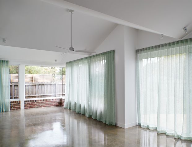 Bright, open-plan interior with polished concrete flooring, white walls and ceiling, floor-to-ceiling windows, and sheer pale green curtains. A ceiling fan hangs over the open space, while glazing opens toward a small enclosed outdoor area with timber fencing and a low brick wall.