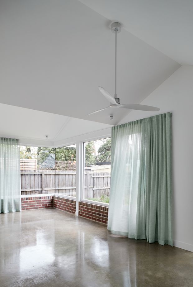 Bright, mostly empty enclosed sunroom with expansive corner windows, pale green sheer and drapery curtains, a white ceiling fan, white walls, exposed red-brick window-height walls, and a polished concrete floor. The room opens visually to a fenced garden area and is filled with soft natural light.