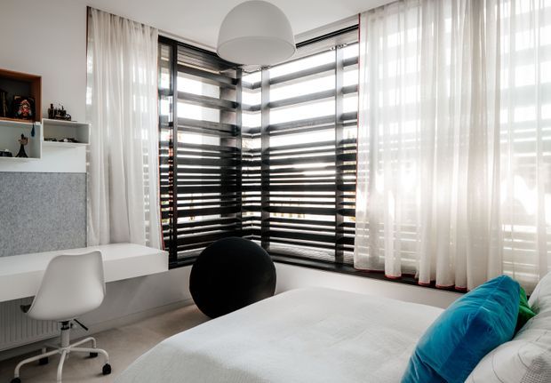 Bright contemporary bedroom with a built-in white study desk and shelving, a large bed with white bedding and vivid blue cushions, and extensive corner glazing fitted with dark horizontal blinds and sheer white curtains. A sculptural white pendant light, black spherical chair, and neutral finishes create a clean, modern interior.