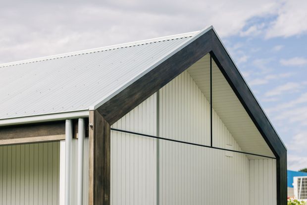 Close-up exterior view of a contemporary gable-front building. The facade combines pale vertical wall cladding with dark timber-like gable framing and a light corrugated metal pitched roof. A covered side opening with slender white posts is visible at left, under a bright overcast sky.