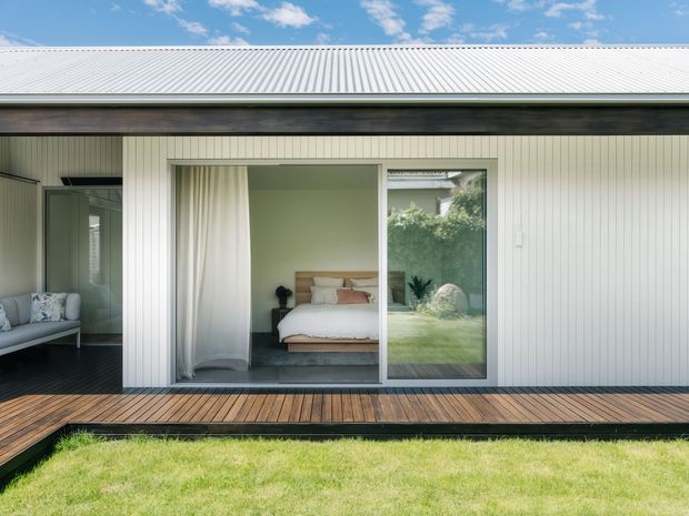 Exterior view of a single-storey contemporary building with a light vertically ribbed metal-clad facade and broad corrugated metal roof. Large glazed sliding doors open onto a narrow timber deck, revealing a simply furnished bedroom inside. The building sits beside a lawn and landscaped garden, under a bright blue sky.