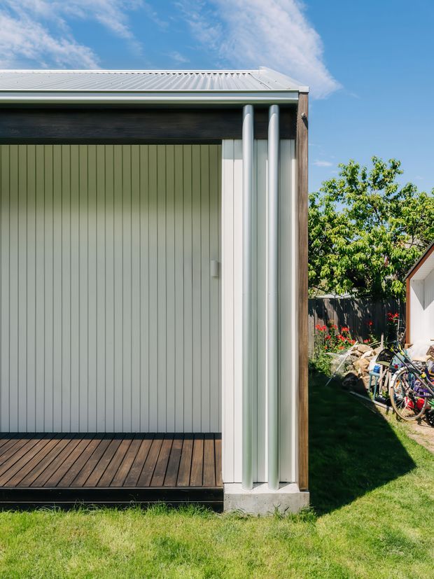 A close exterior view of a compact modern outbuilding or extension with pale vertical wall cladding, a dark horizontal timber feature band, corrugated light-metal roofing, and prominent curved metal downpipes at the corner. A timber deck sits at the base, with a grassed backyard, fencing, trees, flowers, and bicycles visible beside the structure.
