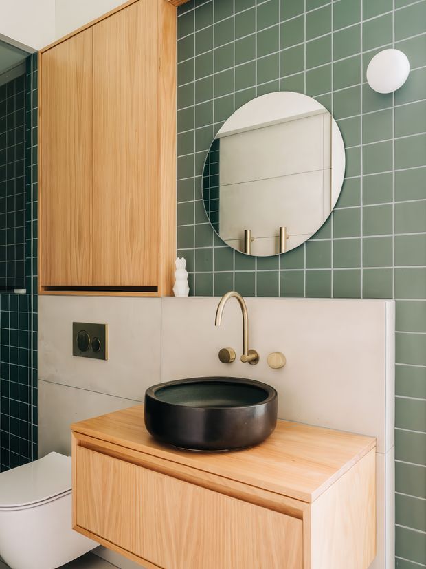 A stylish contemporary bathroom vanity with a wall-mounted timber-grain cabinet, black vessel basin, brushed brass tapware, and a round mirror. The walls combine muted green square tiles with large pale stone-look panels, while a white wall-hung toilet is partially visible at left. The compact space has a refined, warm, modern aesthetic with clean lines and contrasting dark fixtures.