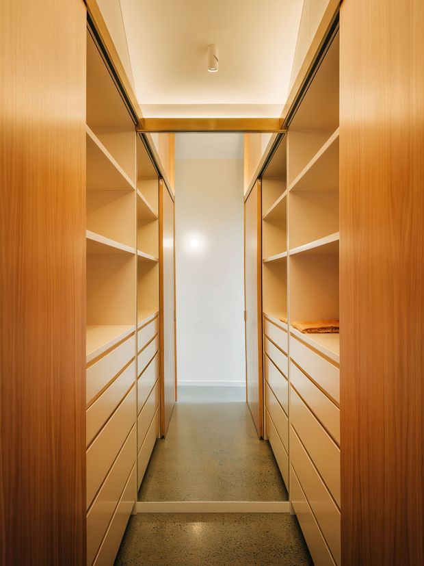 A narrow, walk-in wardrobe or dressing room lined on both sides with full-height timber-toned built-in cabinetry. The cabinetry includes open shelving, multiple shallow drawers, hanging compartments, and a small folded garment. A central doorway leads to a brighter adjoining space, while integrated linear lighting and a warm ceiling light illuminate the room. The floor is a grey polished or speckled concrete-like finish, creating a clean, highly organised, warm contemporary interior.