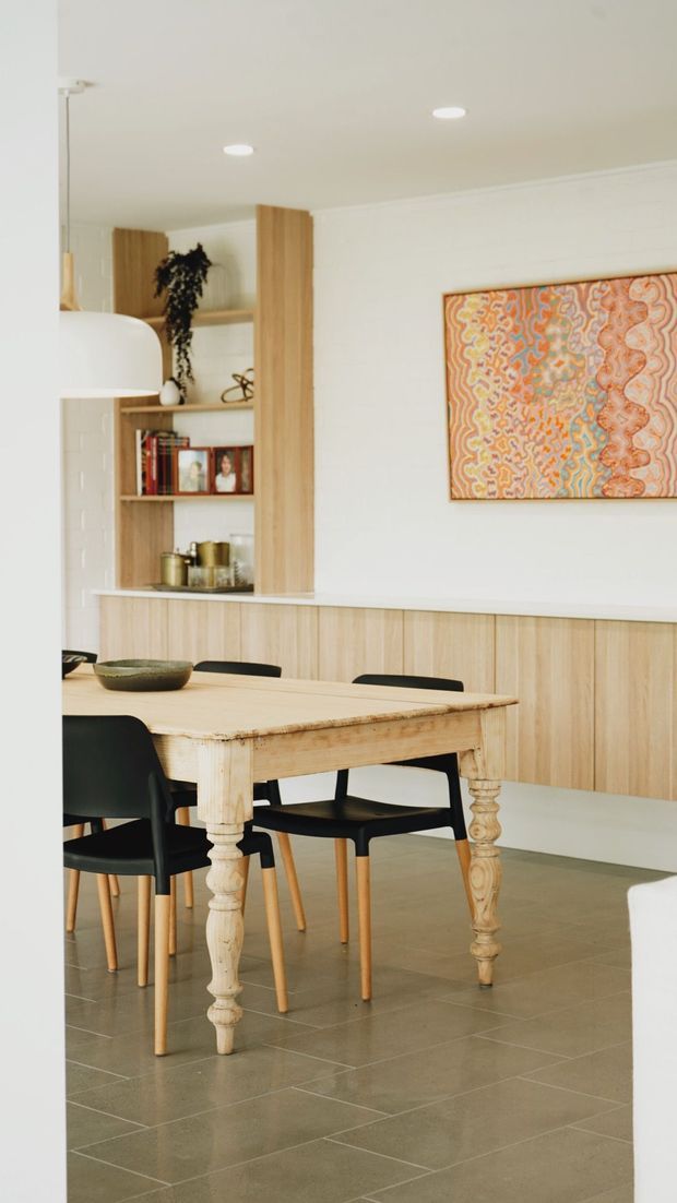 A bright dining area viewed through a doorway, featuring a pale timber dining table with turned legs, black dining chairs, a built-in pale timber display and storage wall, white painted brickwork, recessed ceiling lights, and a colourful framed abstract artwork. The room has a clean, warm contemporary character with restrained natural materials and soft neutral finishes.