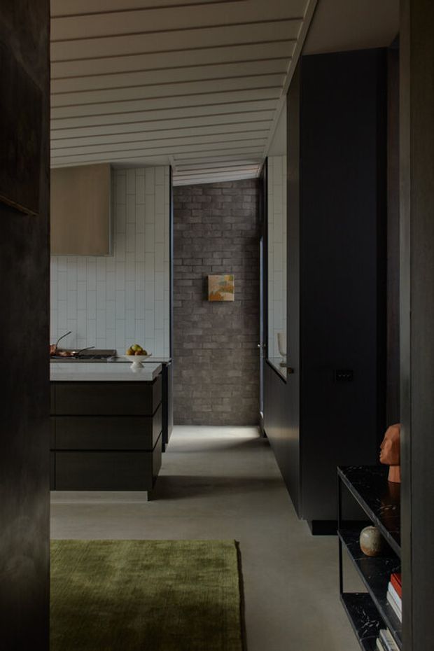 A narrow, moody contemporary interior corridor opening onto a compact kitchen. The space features a dark built-in wall and shelving on the right, charcoal brick feature wall at the rear, pale vertically patterned wall tiles, a dark kitchen island or benchtop with integrated cabinetry, a smooth grey floor, and a white slatted ceiling. A green rug and minimal decorative objects add restrained colour and texture.