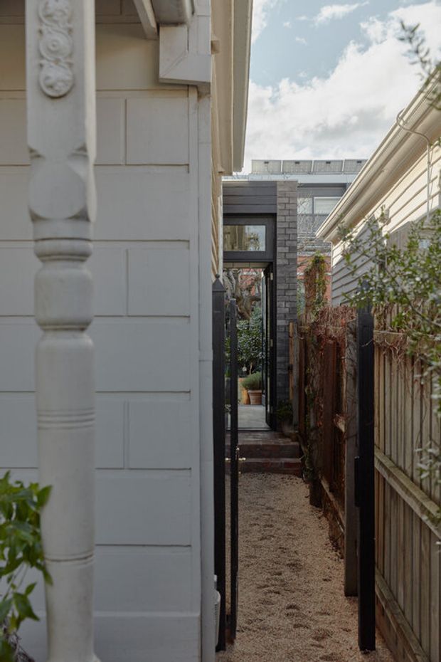 A narrow landscaped passageway between residential structures, with a pale weatherboard wall and ornate timber post on the left, a timber fence and climbing vegetation on the right, and a black-framed glazed doorway leading toward a rear courtyard or garden. Gravel paving and steps create a textured transition space. The image has a restrained heritage-meets-contemporary character under soft daylight.
