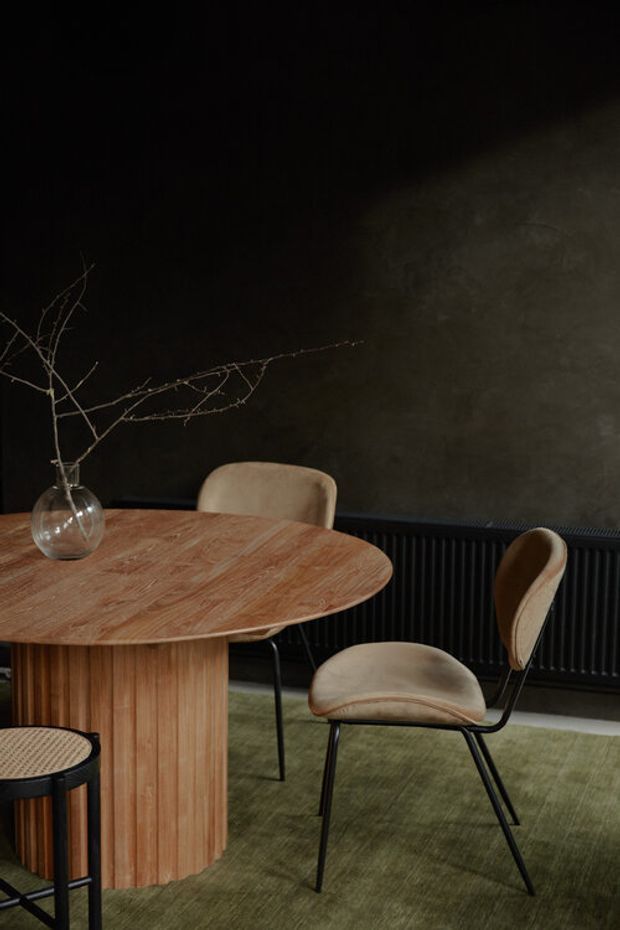 A moody dining area featuring a round light-wood dining table with a fluted pedestal base, upholstered tan dining chairs with black metal frames, and a small woven-seat stool. A clear glass vase with bare branches sits on the table. The room has a dark textured wall, black vertical radiator, and muted green carpet, creating a refined contemporary atmosphere.