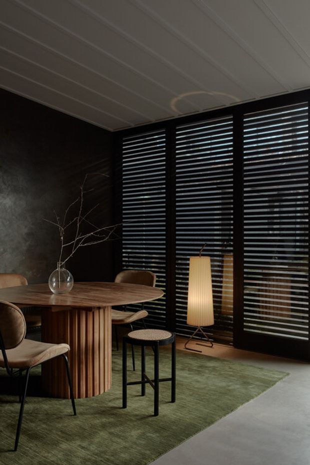 A moody, refined dining room with a round fluted timber dining table and upholstered chairs, set against a dark textured wall. Full-height black-framed louvred glazing lines the back wall, with a sculptural floor lamp, small side table, and muted green rug over a smooth grey floor. The ceiling has white linear panel detailing, creating a strong contrast with the dark walls and glazing.
