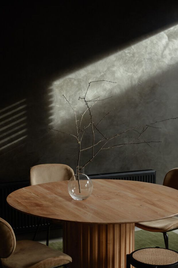 A moody dining room featuring a large round timber dining table with a sculptural fluted pedestal base, surrounded by upholstered dining chairs. A clear glass vase with bare branches forms the centrepiece. The room has dark textured walls, black trim and radiator, dramatic angled sunlight, and a muted green floor covering, creating a refined contemporary atmosphere.