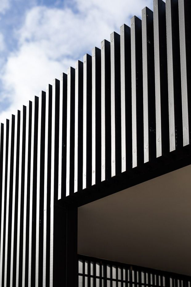 Close-up architectural view of a contemporary black vertical screen or louvred facade, with repeated slender fins rising diagonally against a partly cloudy sky. A dark structural frame and recessed covered opening are visible below, creating strong geometric shadows and contrast.