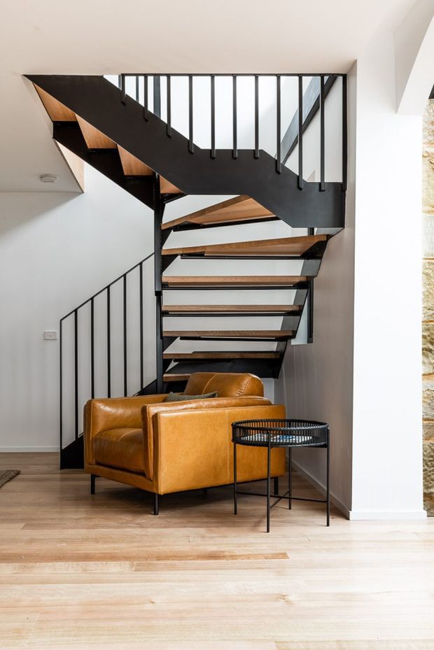 Interior view of a contemporary stairway with open-riser timber treads, black metal stringers and slim vertical balustrades. The staircase occupies a double-height opening beside a warm tan leather armchair and a small round black metal side table. Pale timber flooring and white walls create a bright, refined setting, with a partial exposed stone wall visible at the right edge.