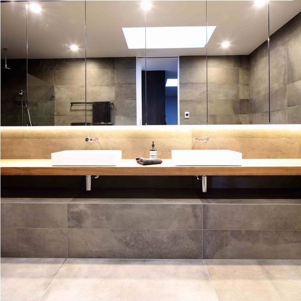Contemporary double-vanity bathroom with large-format grey stone-look tiles on the walls and floor, a timber vanity bench, two white vessel basins, wall-to-wall mirror, under-vanity storage, and warm concealed lighting. A skylight and recessed ceiling lights provide additional illumination, while a shower area is visible in the mirror reflection.