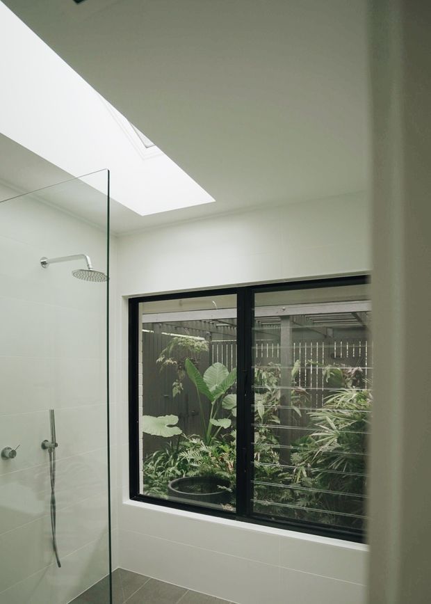 Bright contemporary bathroom with a frameless glass walk-in shower, wall-mounted rain shower and handheld fixture, large white wall tiles, grey tiled floor, and a black-framed window overlooking a planted outdoor area. A skylight brings in additional natural light.