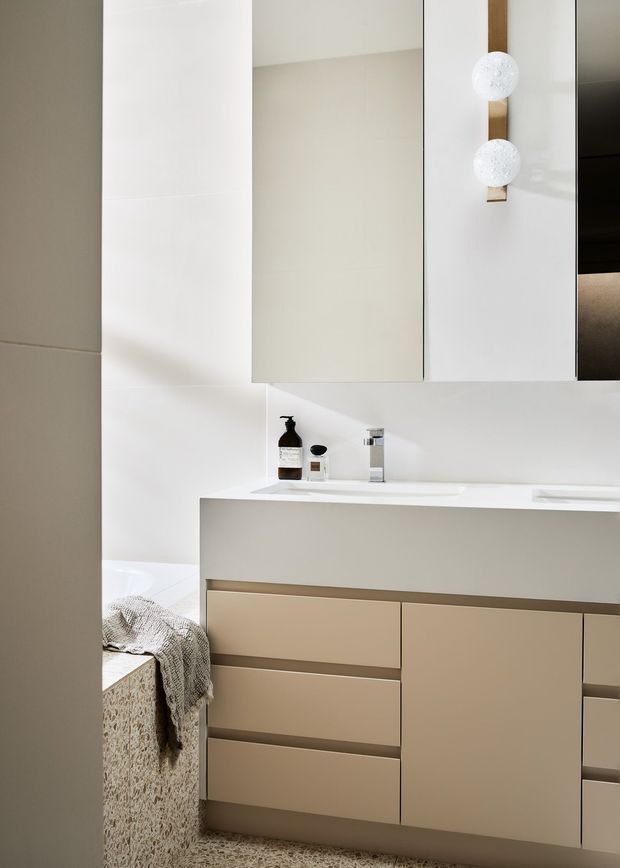 A contemporary minimalist bathroom vanity with a long white integrated basin and pale taupe cabinetry. The room features large white wall surfaces, mirrored upper cabinets, a chrome tap, and a vertical brass-toned light fitting with two spherical white globes. A freestanding or partially visible bathtub with a textured draped towel appears at the left edge.