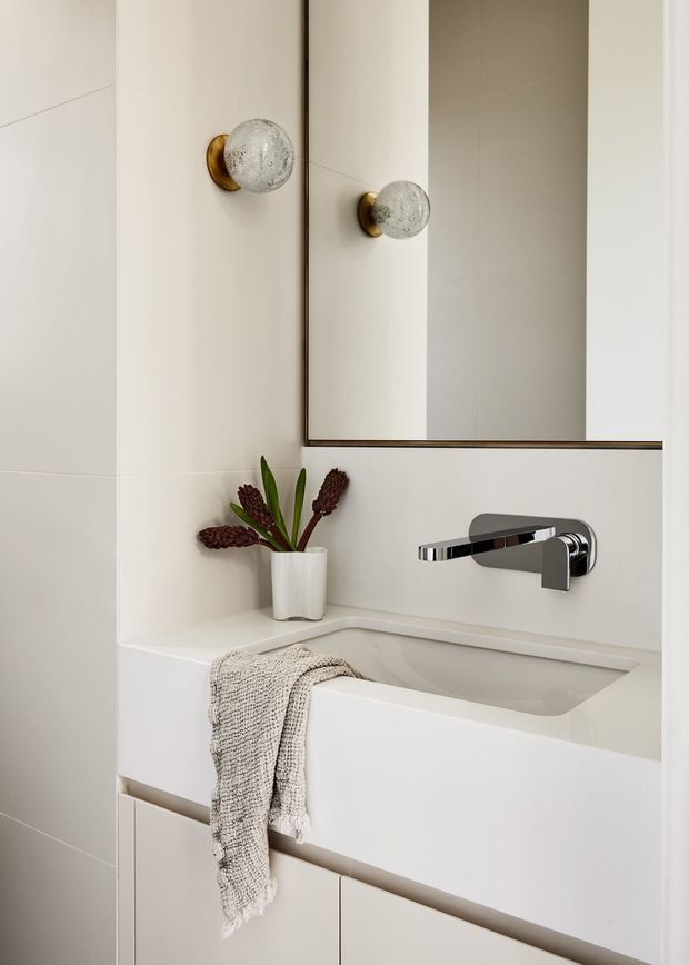 A compact, refined bathroom vanity featuring a white integrated rectangular basin, wall-mounted chrome tapware, pale large-format tiled walls, and white fixed cabinetry. A frameless or slim dark-edged mirror spans the vanity, with decorative brass-and-glass wall sconces on either side. Neutral styling is complemented by a small vase with dried botanicals and a textured beige towel.