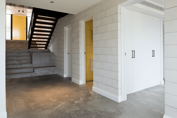 Interior view of a modern entry hall with a raw concrete floor, white painted block walls, integrated white storage cabinetry, and a split-level staircase. The staircase combines concrete steps and a dark steel structure, with a yellow door and matching yellow glazed panel adding a strong accent.