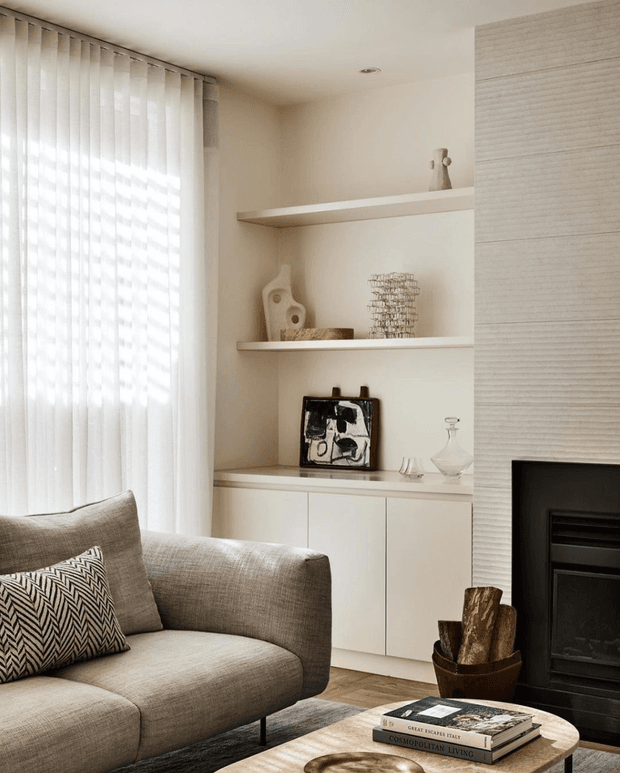 A softly lit, neutral-toned living room featuring a large upholstered sofa, sheer vertical window curtains, built-in white display shelving and cabinetry, and a textured feature wall surrounding a black fireplace. Minimal sculptural decor, timber flooring, and a round timber coffee table create a calm, refined interior.
