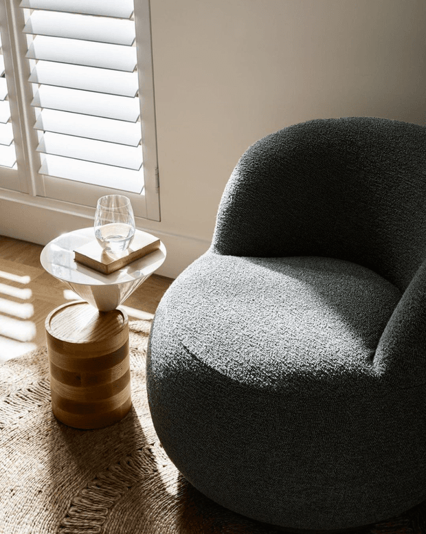 A softly lit interior vignette featuring a large rounded charcoal boucle lounge chair beside a small sculptural glass-topped side table and cylindrical timber stool. White plantation shutters filter strong sunlight across a warm timber floor and natural woven rug. A drinking glass and book add a quiet, refined living-room setting.