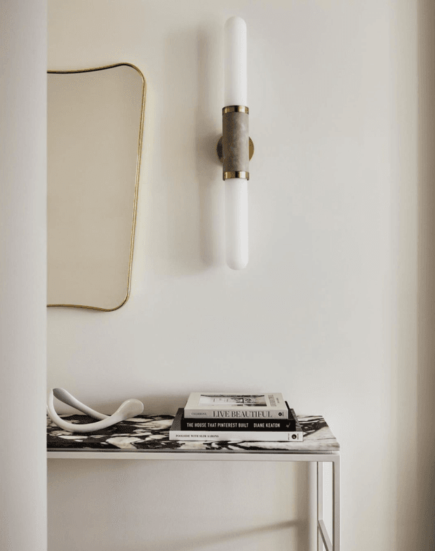 A close-up interior vignette featuring a slim white wall sconce with brass and dark stone or metal detailing, mounted above a narrow console table. The console has a striking black-and-white patterned stone-like top and slender light-coloured legs, styled with stacked design books and a sculptural white object. The overall composition is softly lit, neutral, refined, and minimalist with subtle luxe detailing.