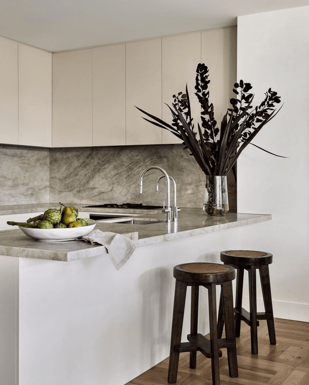 A refined contemporary kitchen with a large white island, integrated cooktop and sink, pale full-height cabinetry, and a textured grey-beige stone splashback and benchtop. Two dark timber stools sit at the island, while a sculptural arrangement of dark foliage provides a dramatic accent. Timber flooring adds warmth to the otherwise neutral space.