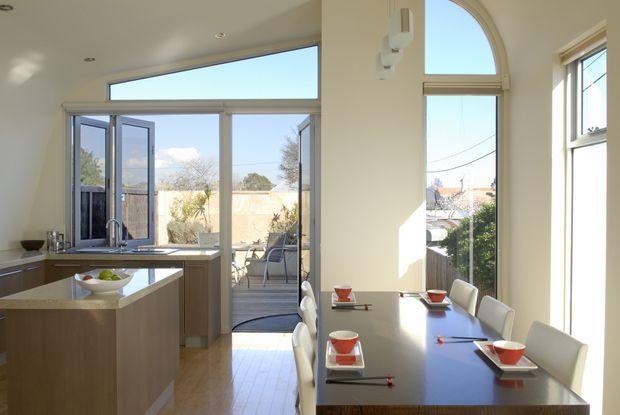 Bright open-plan kitchen and dining area with a compact central island, pale timber cabinetry, light benchtops, and extensive glazing opening to a small deck or patio. The dining zone features a long dark table with white upholstered chairs, while tall and arched windows bring in abundant natural light and views toward the surrounding residential landscape.