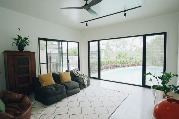 Bright contemporary living room with white walls and ceiling, large black-framed sliding glass doors overlooking a swimming pool and landscaped garden, a dark sectional sofa with mustard cushions, patterned rug, timber cabinet, ceiling fan, track lighting, and indoor plants.
