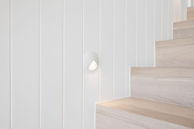 Minimalist interior stairway with pale timber treads and risers beside a white vertical wall-panel feature. A small sculptural wall light provides a subtle warm accent.
