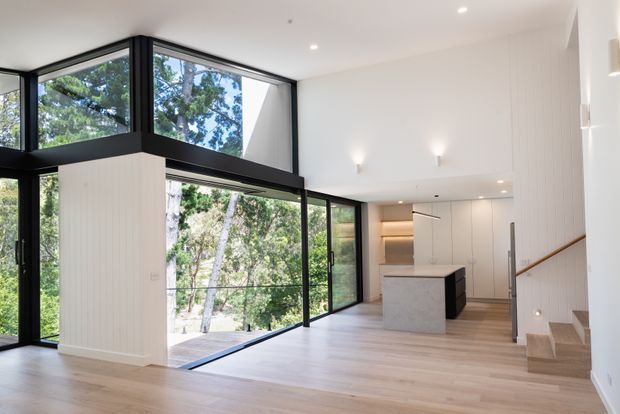 Bright, open-plan contemporary interior with a kitchen and island, pale timber flooring, white vertical wall panelling, and extensive black-framed floor-to-ceiling glazing overlooking trees. A stairway with timber handrail and open-riser detailing is visible at right, while recessed lighting and minimalist wall sconces contribute to the clean architectural character.