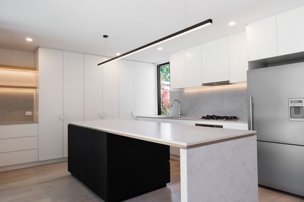Contemporary kitchen with a large white island featuring black cabinetry, a pale stone-look benchtop and waterfall end panel. White full-height cabinetry, integrated appliances, a stainless-steel refrigerator, gas cooktop, light timber flooring, and a slim linear pendant create a clean, high-contrast interior.