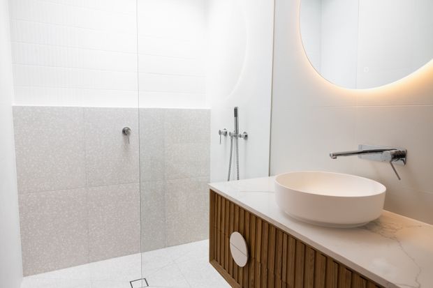 Bright contemporary bathroom with a frameless glass shower, white tiled walls, pale terrazzo-look wall tiles, a round vessel basin, and a floating timber-slat vanity with integrated storage. A large rounded mirror with concealed backlighting adds a soft glow.